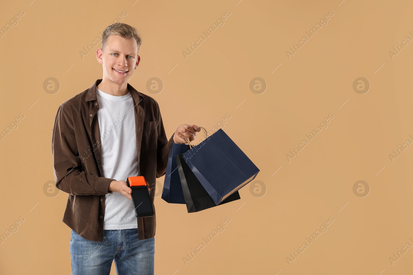 Photo of Happy young man with payment terminal and shopping bags on beige background, space for text