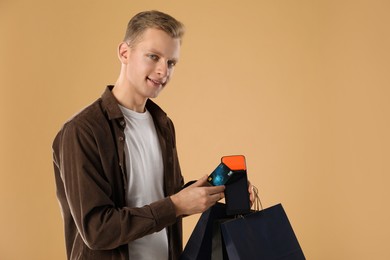 Happy young man with payment terminal, shopping bags and debit card on beige background