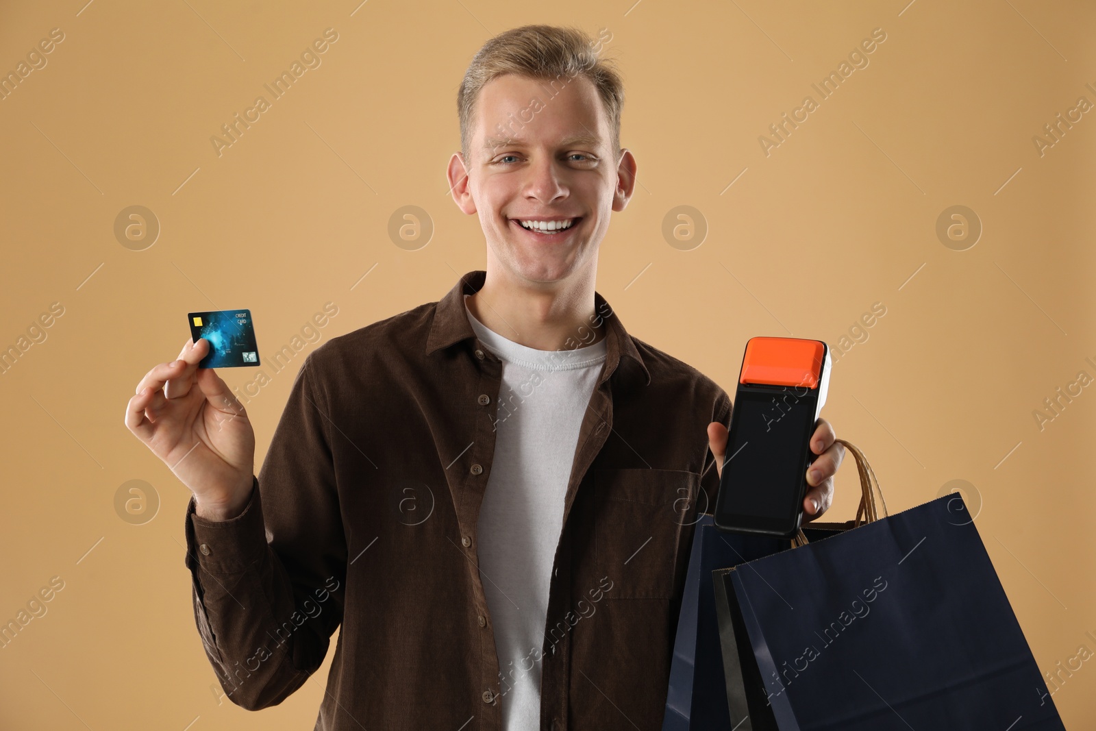 Photo of Happy young man with payment terminal, shopping bags and debit card on beige background