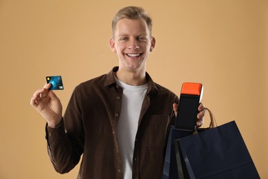 Happy young man with payment terminal, shopping bags and debit card on beige background