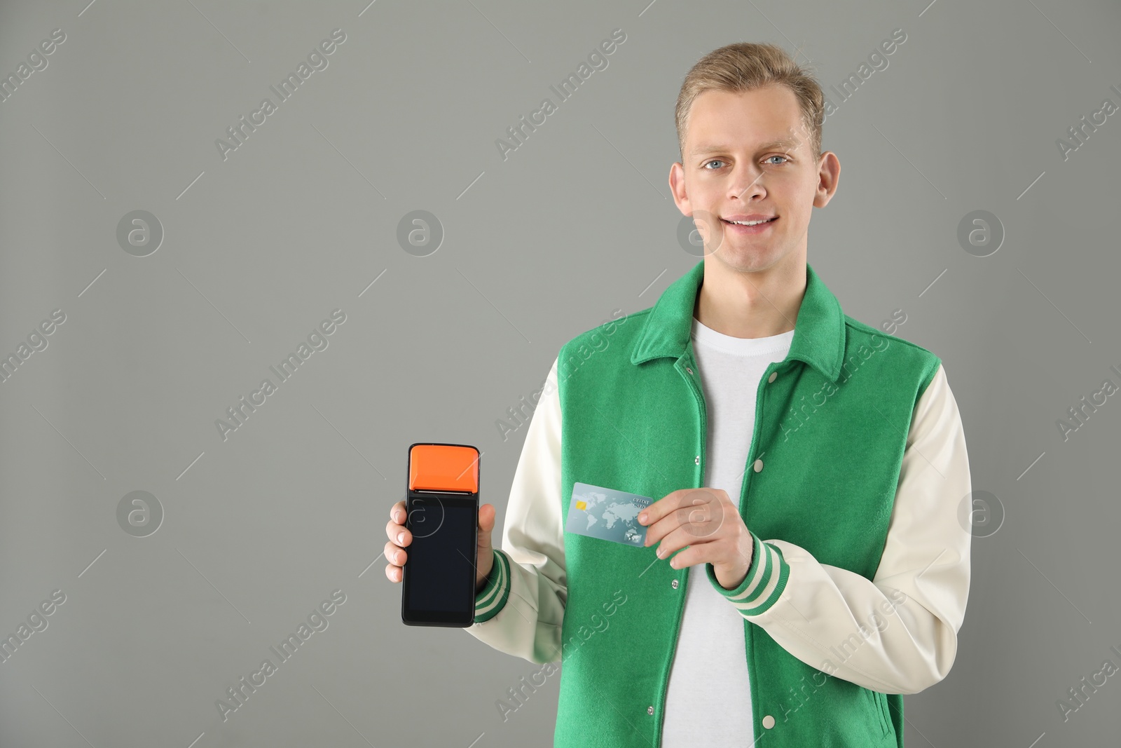 Photo of Happy young man with payment terminal and debit card on light grey background, space for text