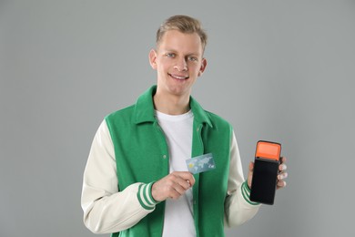 Happy young man with payment terminal and debit card on light grey background