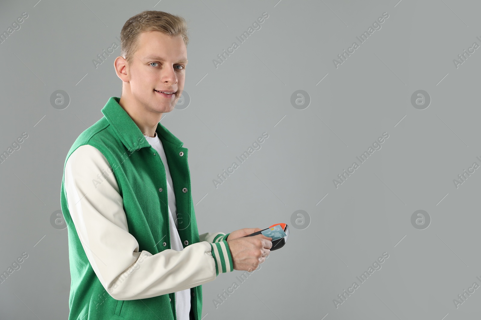 Photo of Happy young man with payment terminal and debit card on light grey background, space for text