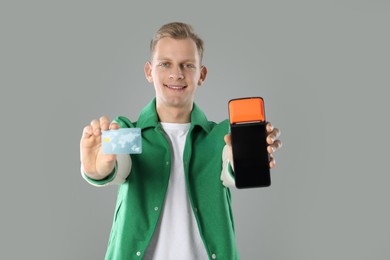 Happy young man with payment terminal and debit card on light grey background