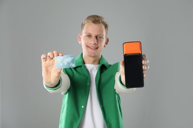 Happy young man with payment terminal and debit card on light grey background, selective focus