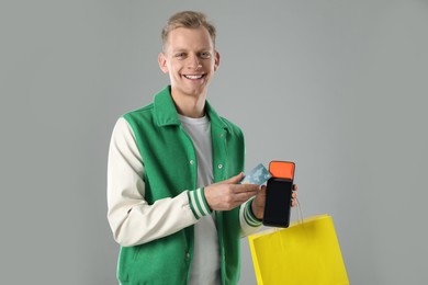 Happy young man with payment terminal, shopping bag and debit card on light grey background