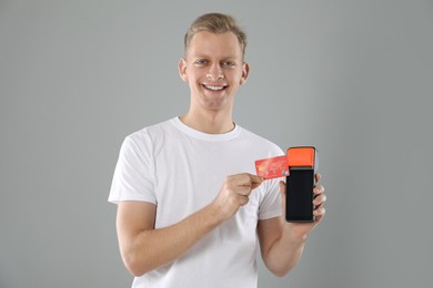 Photo of Happy young man with payment terminal and debit card on light grey background