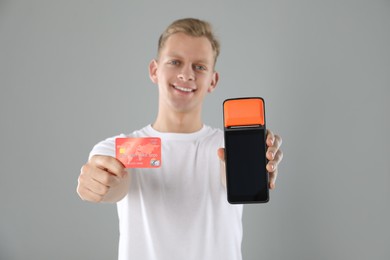 Happy young man with payment terminal and debit card on light grey background, selective focus