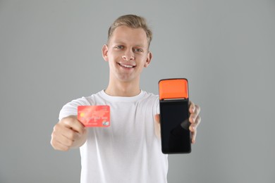 Happy young man with payment terminal and debit card on light grey background