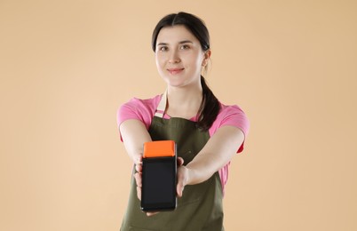 Photo of Young woman in apron with payment terminal on beige background