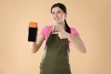 Happy young woman in apron with payment terminal on beige background