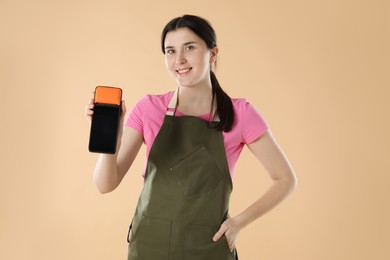 Happy young woman in apron with payment terminal on beige background