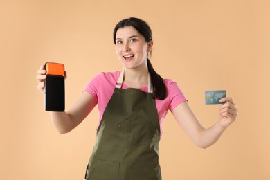 Photo of Happy young woman in apron with payment terminal and debit card on beige background