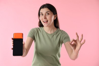 Photo of Happy young woman with payment terminal showing OK gesture on pink background