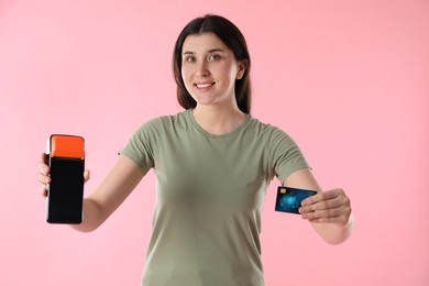 Happy young woman with payment terminal and debit card on pink background