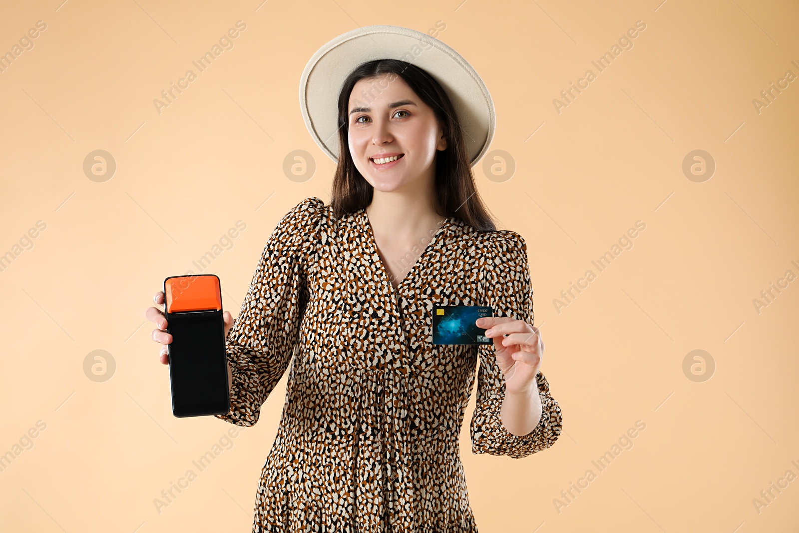 Photo of Happy young woman with payment terminal and debit card on beige background