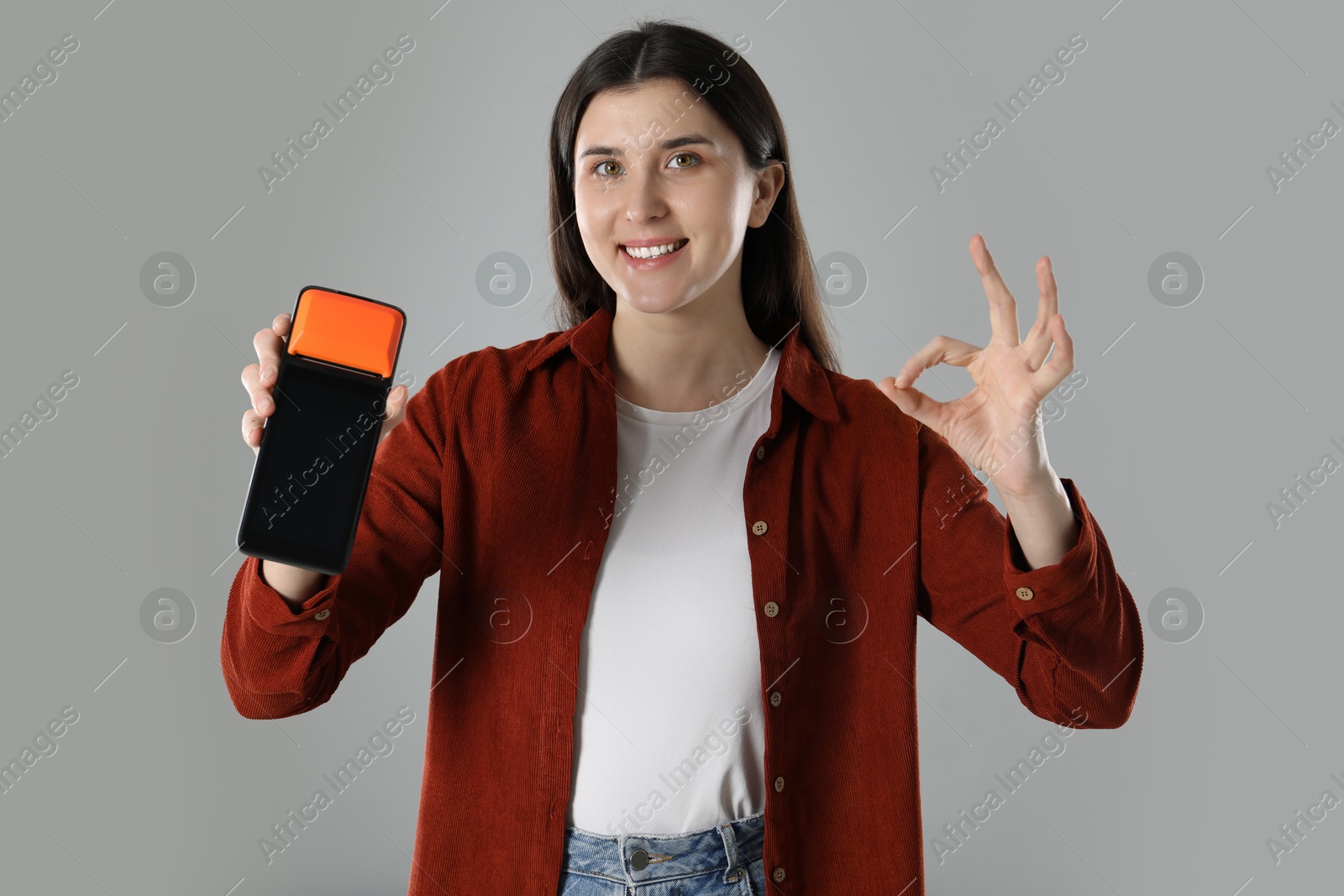 Photo of Happy young woman with payment terminal showing OK gesture on gray background