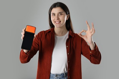 Happy young woman with payment terminal showing OK gesture on gray background