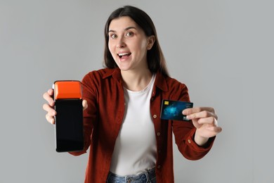 Photo of Surprised young woman with payment terminal and debit card on gray background