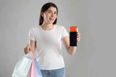 Happy young woman with payment terminal and shopping bags on gray background, space for text