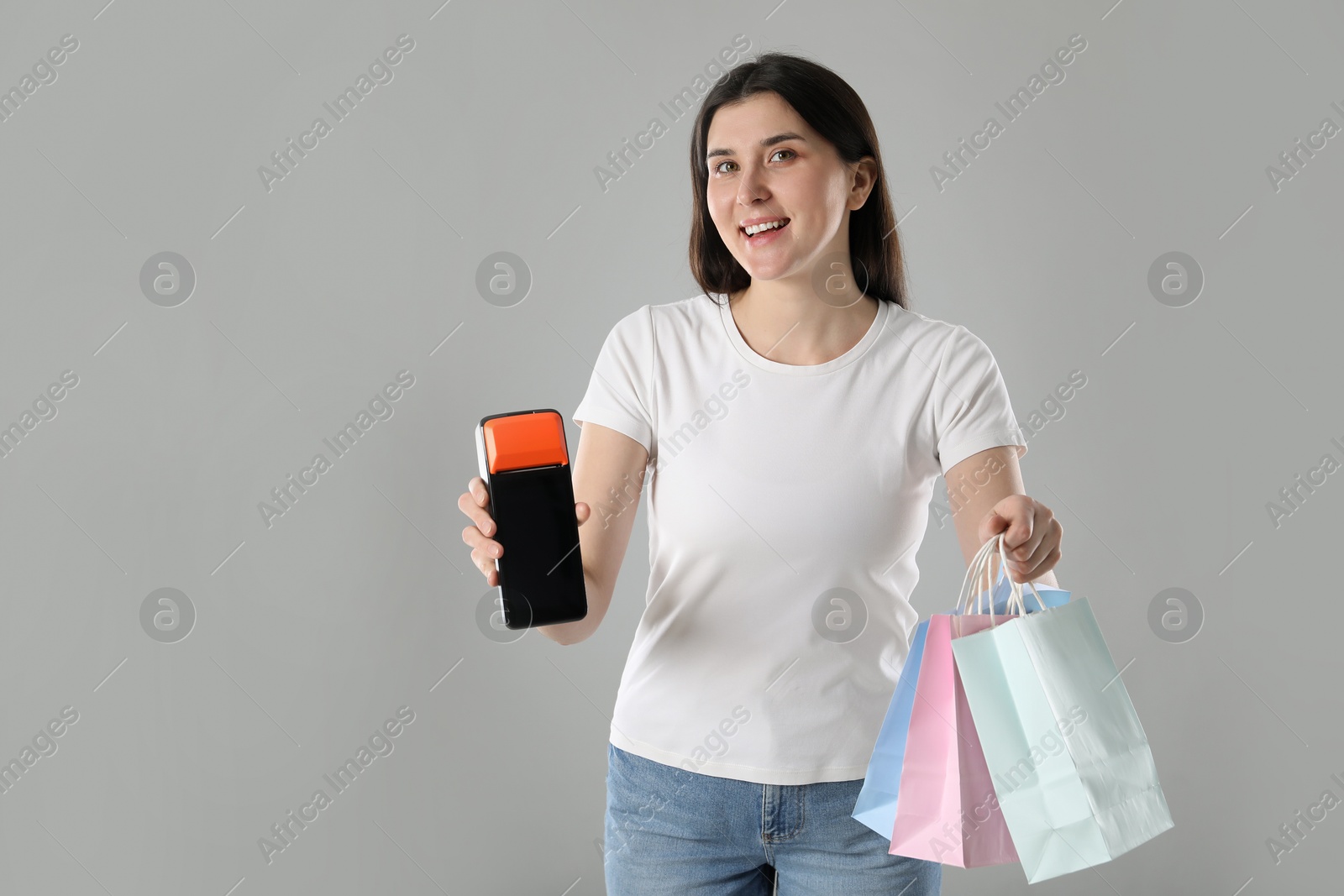 Photo of Happy young woman with payment terminal and shopping bags on gray background, space for text