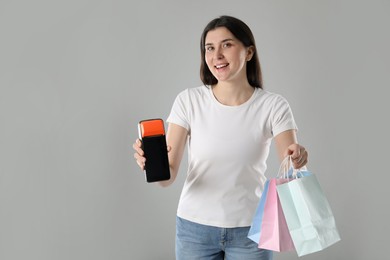 Happy young woman with payment terminal and shopping bags on gray background, space for text