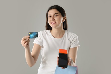 Photo of Happy young woman with payment terminal, shopping bags and debit card on gray background