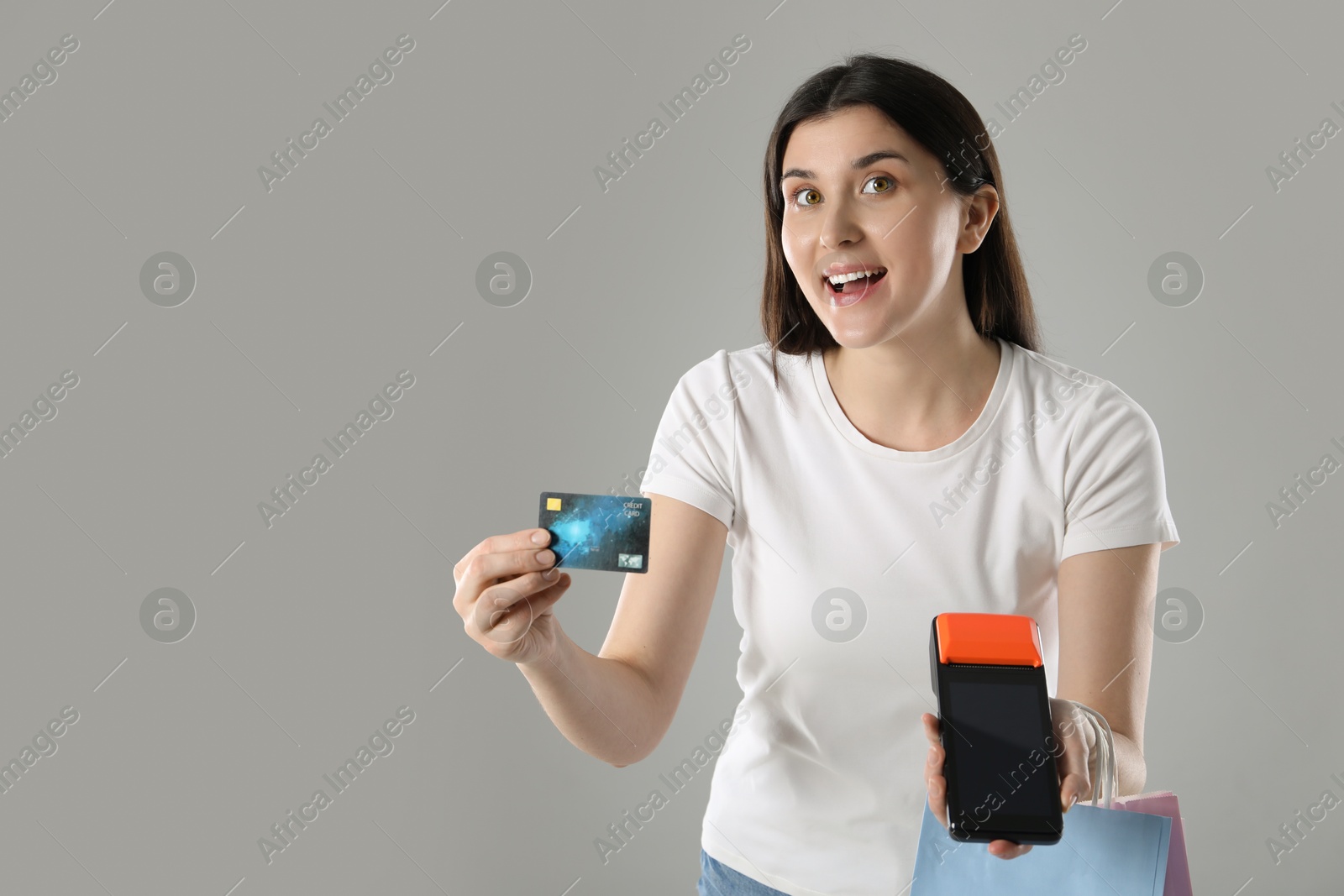 Photo of Happy young woman with payment terminal and shopping bags on gray background, space for text