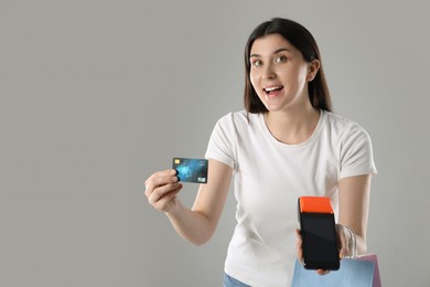 Happy young woman with payment terminal and shopping bags on gray background, space for text