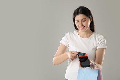 Photo of Happy young woman with payment terminal, shopping bags and debit card on gray background, space for text