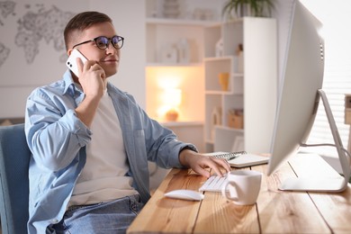 Photo of Handsome man talking by smartphone at desk with computer indoors. Home office