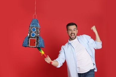 Photo of Emotional man hitting rocket shaped pinata with stick on red background