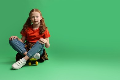 Photo of Stylish little girl sitting on penny board against green background, space for text