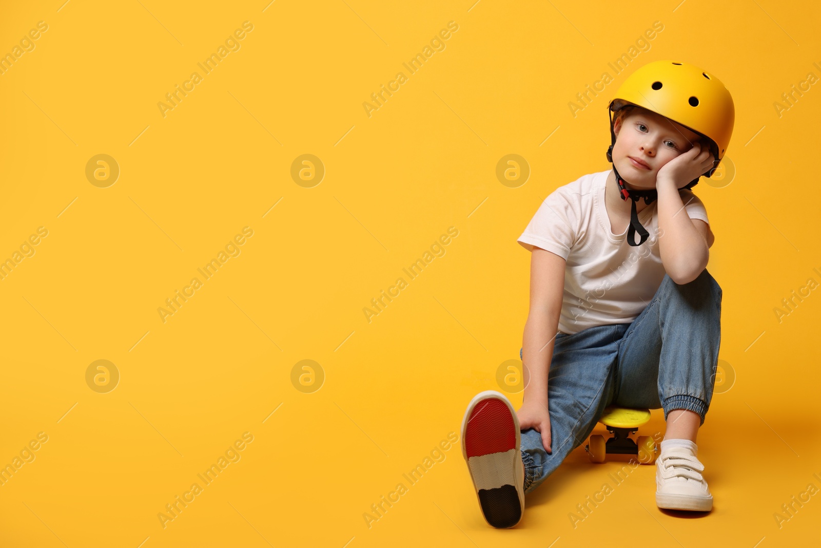 Photo of Little girl in helmet sitting on penny board against orange background, space for text