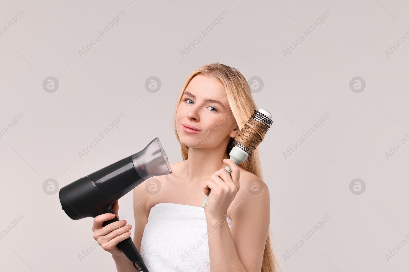 Photo of Beautiful young woman styling her hair with hairdryer and brush on light grey background