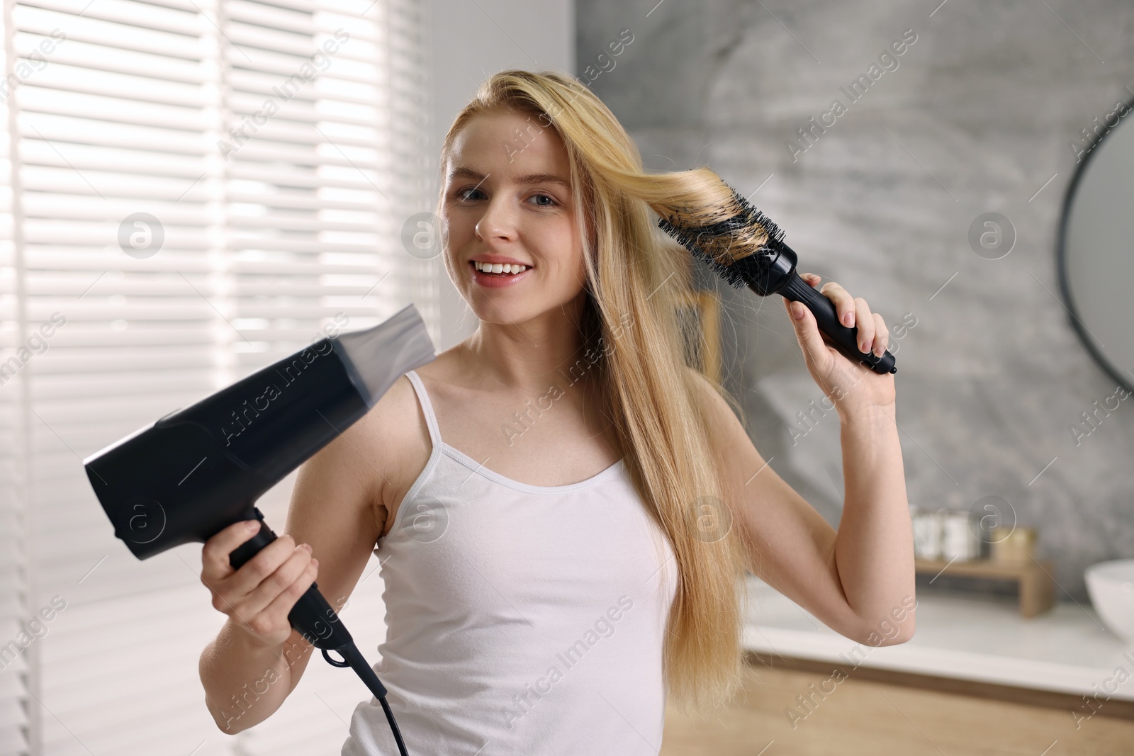 Photo of Beautiful young woman styling her hair with hairdryer and brush in bathroom