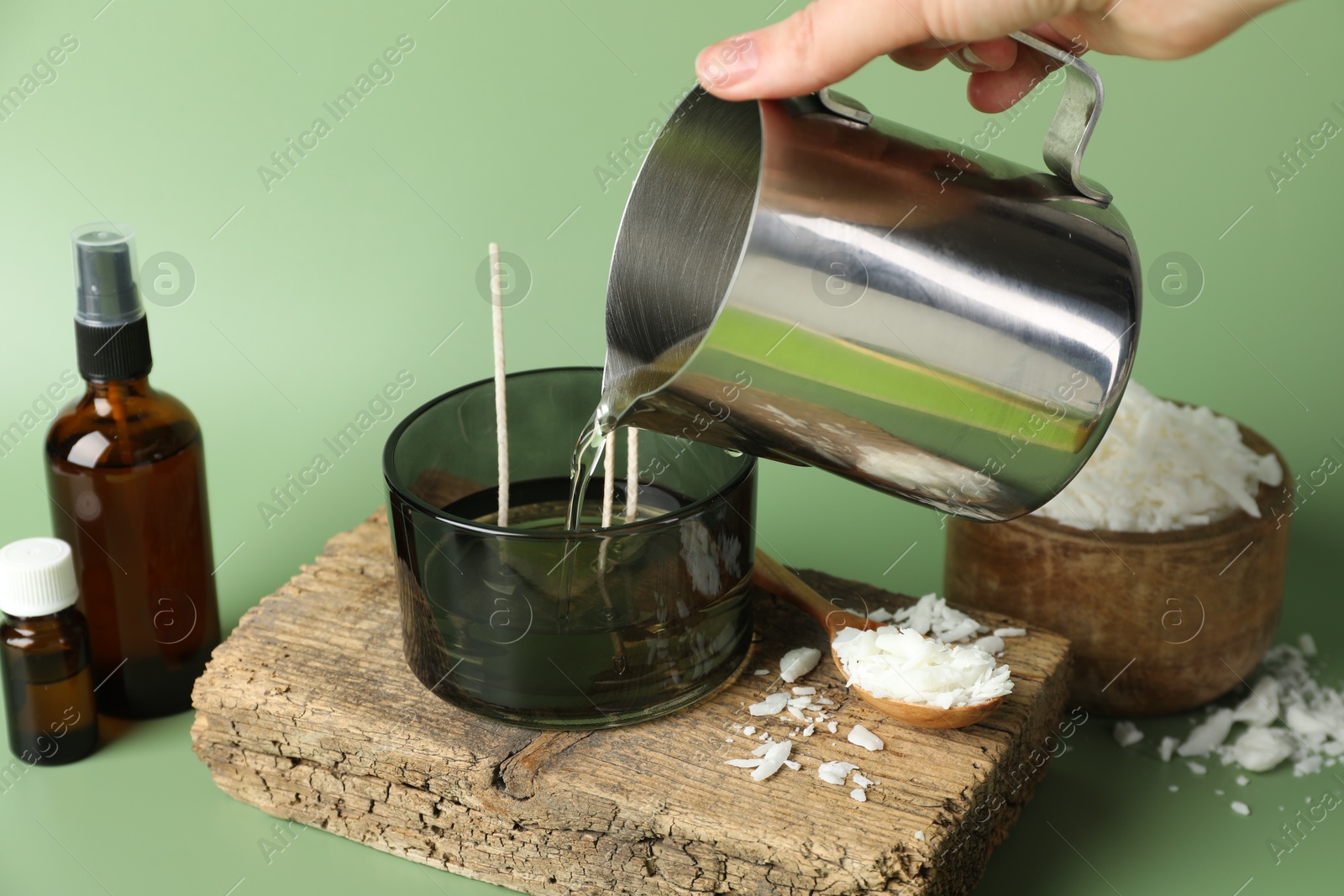 Photo of Making handmade candles. Woman pouring melted soy wax into jar on green background, closeup