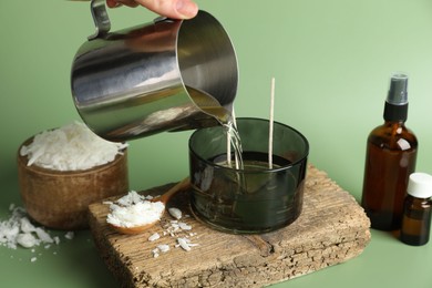 Photo of Making handmade candles. Woman pouring melted soy wax into jar on green background, closeup