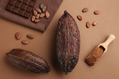 Cocoa pods, scoop with powder, beans and chocolate bar on brown background, flat lay