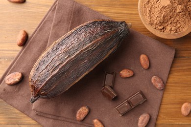 Photo of Cocoa pod, powder in bowl, beans and chocolate pieces on wooden table, flat lay