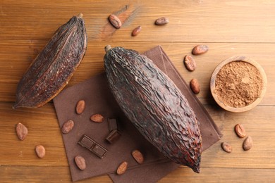 Photo of Cocoa pods, powder in bowl, beans and chocolate pieces on wooden table, flat lay