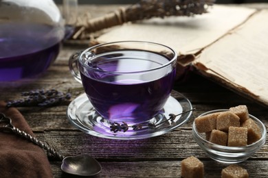 Photo of Aromatic lavender tea in glass cup, brown sugar, book, spoon and dry flowers on wooden table, closeup