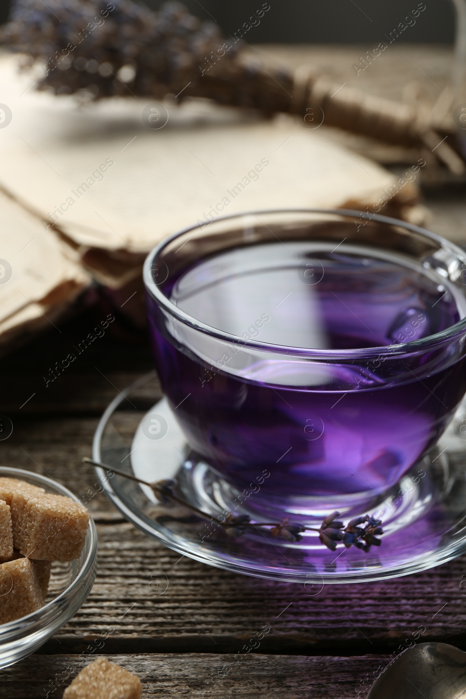 Photo of Aromatic lavender tea in glass cup, brown sugar, book and dry flowers on wooden table, closeup