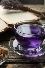 Photo of Aromatic lavender tea in glass cup, brown sugar, book and dry flowers on wooden table, closeup