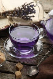 Photo of Aromatic lavender tea in glass cup, brown sugar, book, spoon and dry flowers on wooden table, closeup