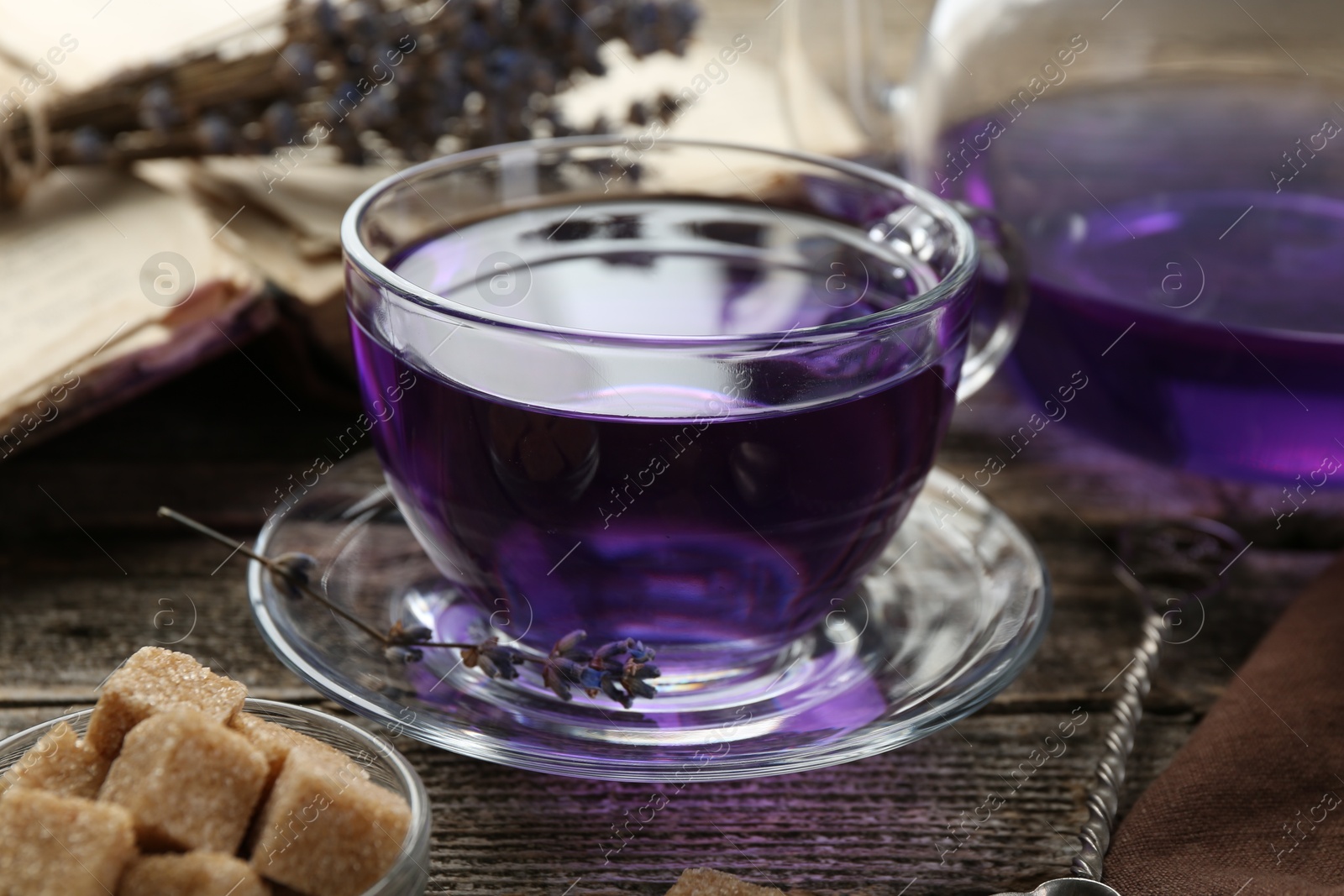 Photo of Aromatic lavender tea in glass cup, brown sugar, book and dry flowers on wooden table, closeup