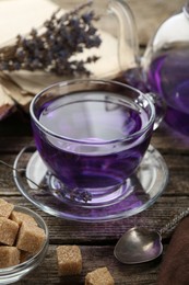 Photo of Aromatic lavender tea in glass cup, spoon, brown sugar, teapot, book and dry flowers on wooden table, closeup