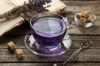 Photo of Aromatic lavender tea in glass cup, brown sugar, book, spoon and bunch of dry flowers on wooden table, closeup