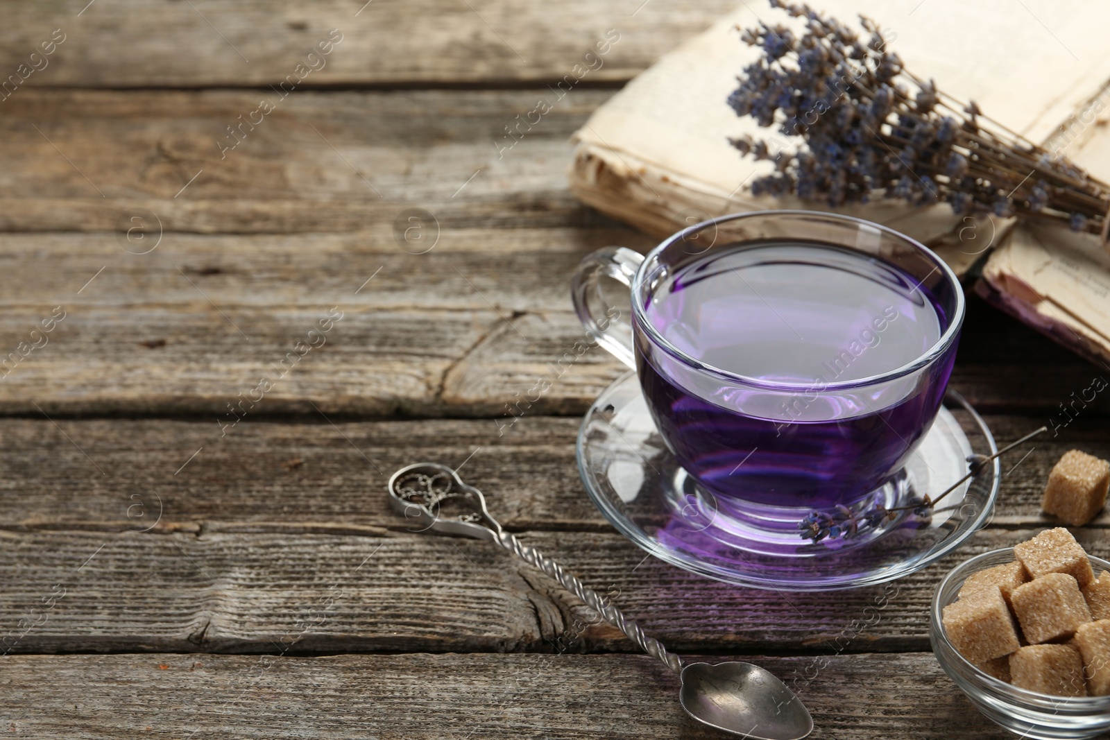 Photo of Aromatic lavender tea in glass cup, brown sugar, book, spoon and bunch of dry flowers on wooden table, closeup. Space for text