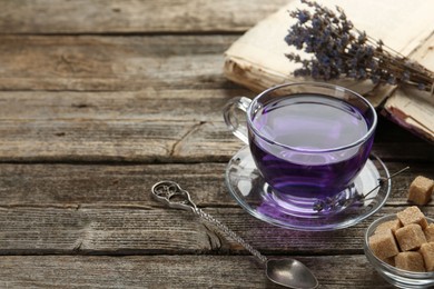 Photo of Aromatic lavender tea in glass cup, brown sugar, book, spoon and bunch of dry flowers on wooden table, closeup. Space for text